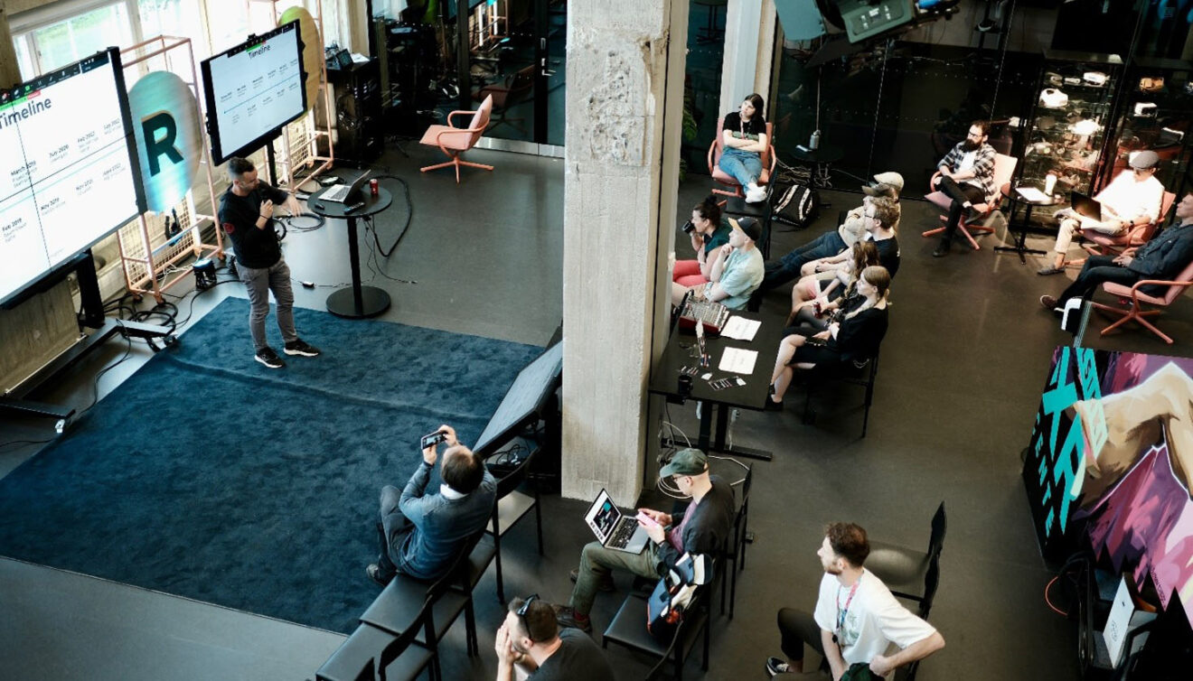 Overhead view of a small audience watching a speaker present slides in an industrial-style event space.
