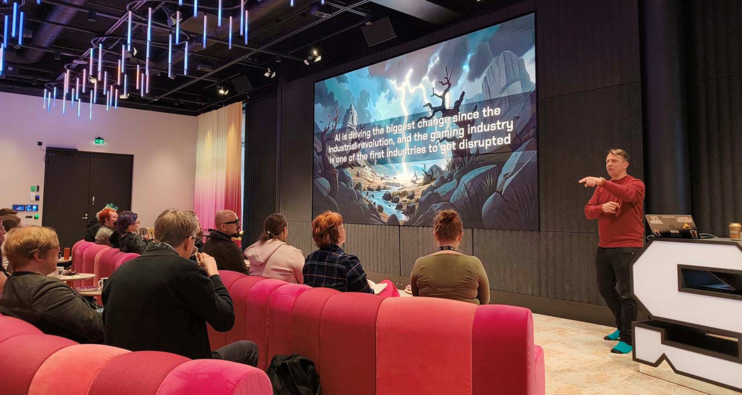 Front row view of people listening to a talk in an auditorium. Slide presented includes cartoonish visuals and states “AI is driving the biggest change since the industrial revolution, and the gaming industry is one of the first industries to get disrupted.