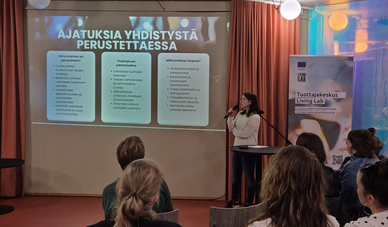 Puhuja esittelee yleisölle diaa ”Ajatuksia yhdistystä perustettaessa”, oikealla näkyy Tuottajakeskus Living Lab -rollup.